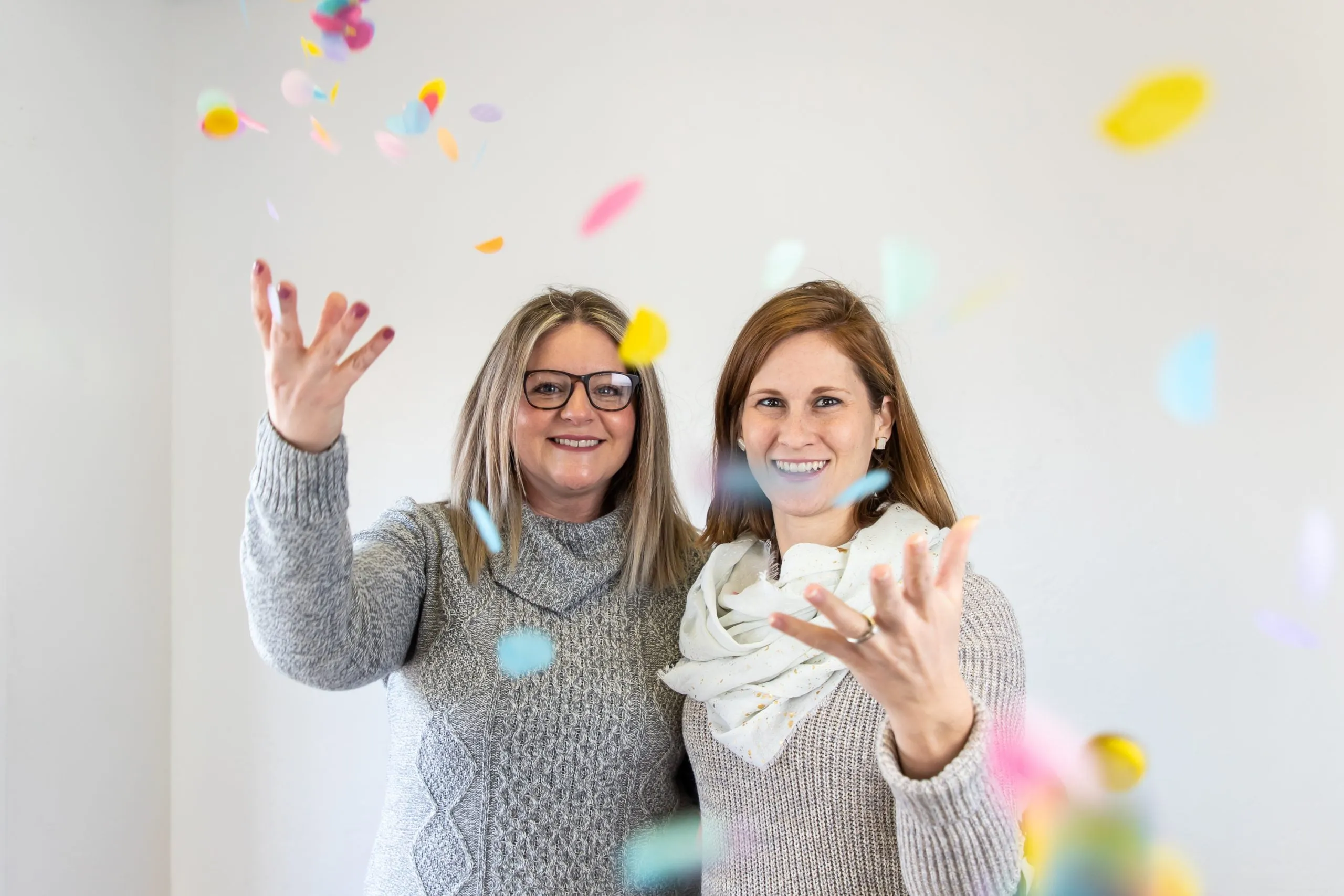 Two women smiling while throwing colorful confetti in the air. They both are wear sweaters. 