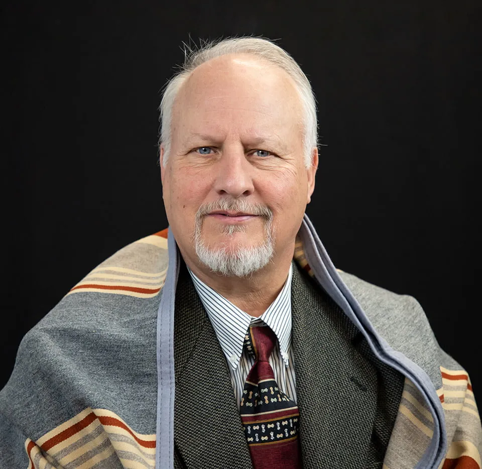 An older man is photographed on a black background wearing a layered suit. He has a patterned blanket covering his shoulders. 
