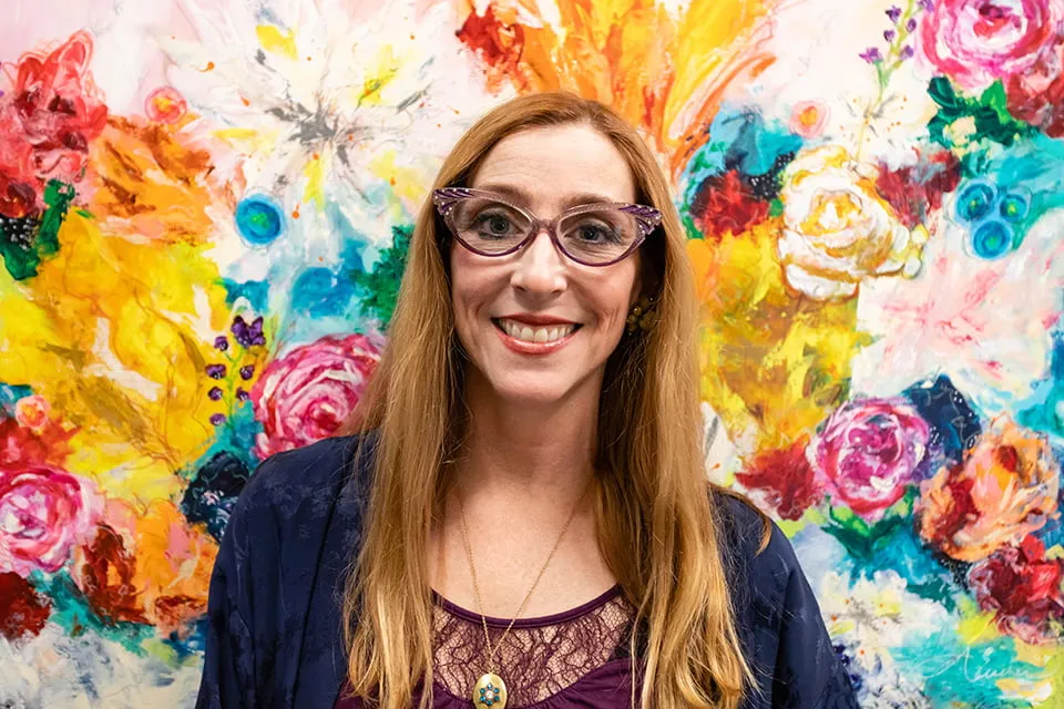 Hope from Ash - myheartcreative Aimee Eischen smiles for a portrait. She has red hair and purple glasses. The background is a large, colorful floral painting created by her.