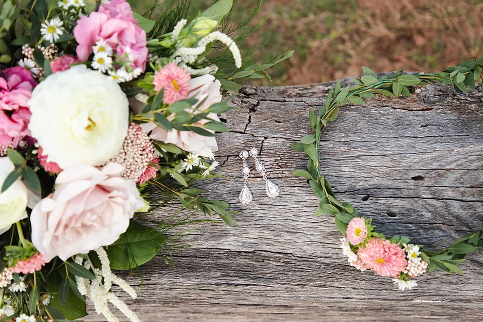 A pair of diamond earrings, a flower crown, and a bouquet of pink and white flowers rest on a log. 