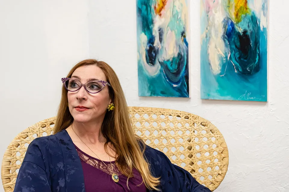 Hope from Ash - myheartcreative Aimee Eischen sits in a wicker chair and gazes to the right. She is photographed sitting in front of two abstract, blue paintings hanging on the wall.