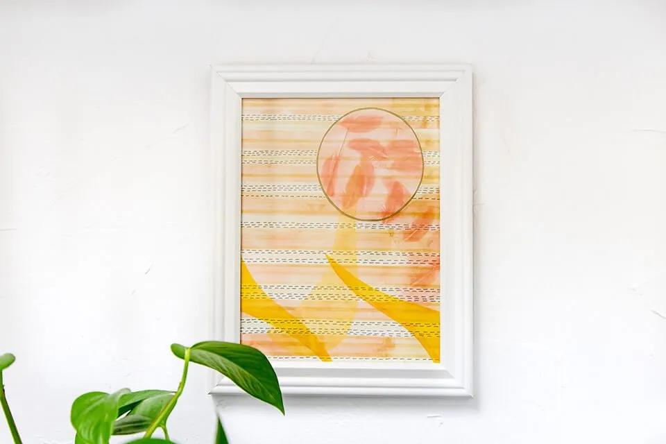 An abstract piece by Emma Difani hanging on the myheartcreative studio wall. There is a green plant sitting below it. The piece has light pink, yellow, and orange tones. 