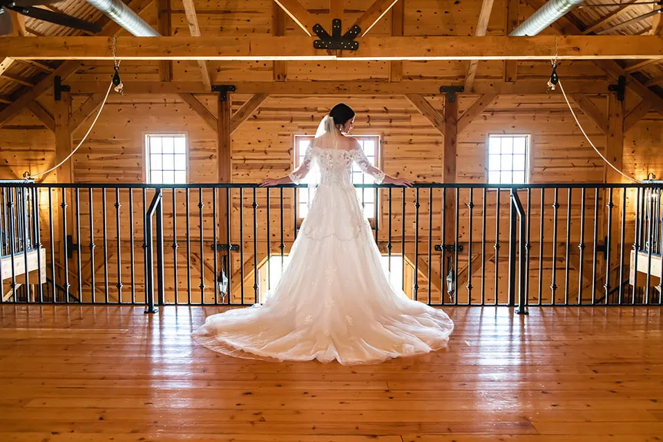 The bride holds on to the upstairs railing while overlooking the downstairs floor of McGranahan Barn. Her dress is on display. 