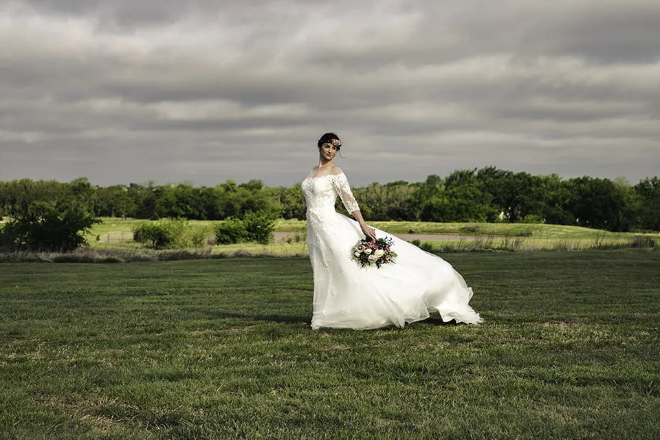A bride wears a wedding gown from Bella Rose Bridal. She stands in a field as the wind blows her dress. 