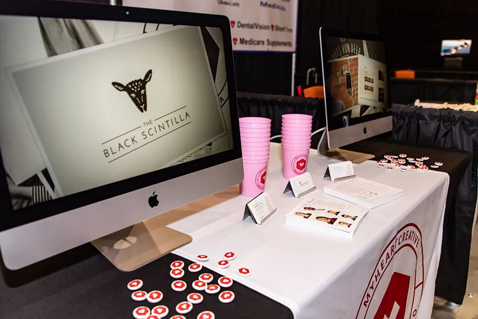 Chahtapreneurs - myheartcreative The myheartcreative booth at the Choctaw Summit and Business Far. The table has a pile of myheartcreative buttons, pink branded cups, business cards and table cards, and two Mac desktops with slideshows of client work.