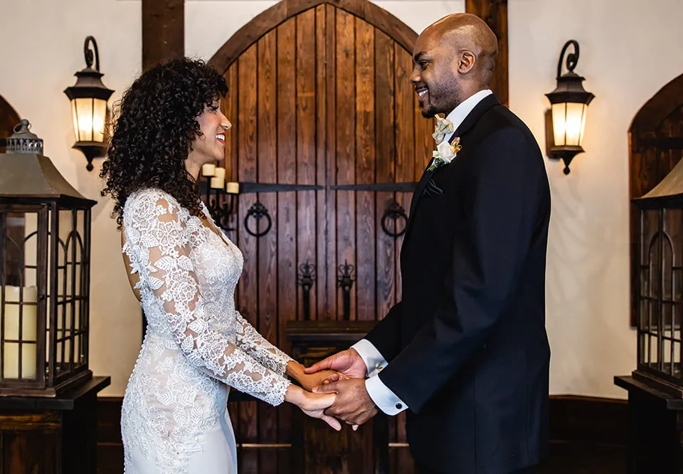 The bride and groom hold hands and smile at one another as they stand in front of large wooden doors at Rose Briar Place. 