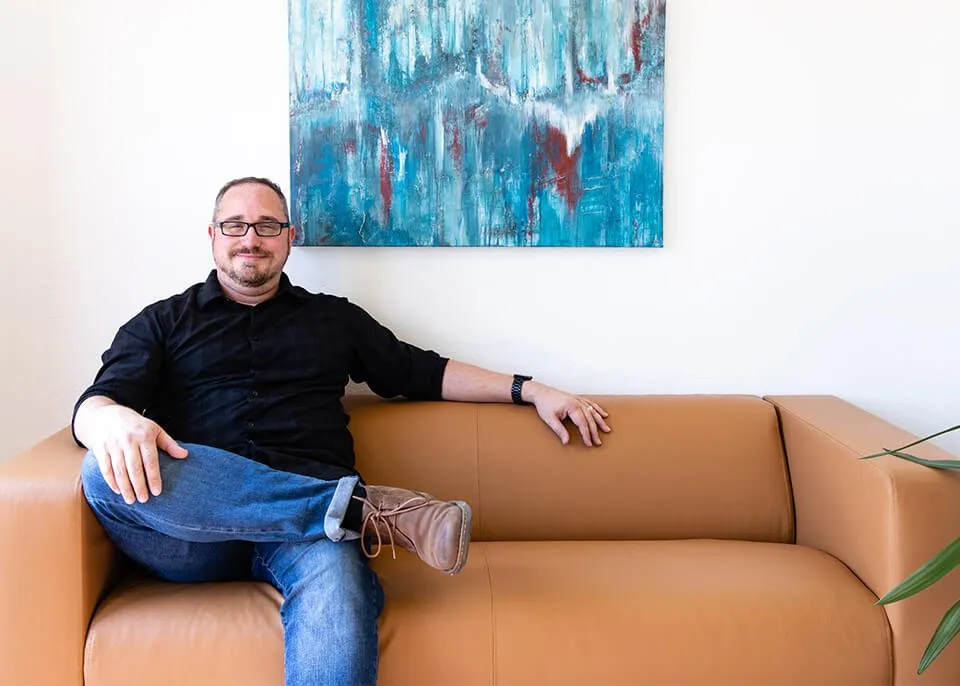 Artist Keith Wolfe sits on a tan sofa at the myheartcreative studio. One of his abstract paintings hangs on the wall behind him. 