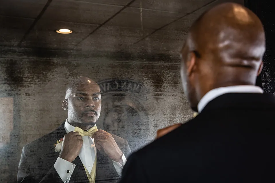 The groom adjusts his gold bow tie as he looks in the mirror in the Grooms Suite. 