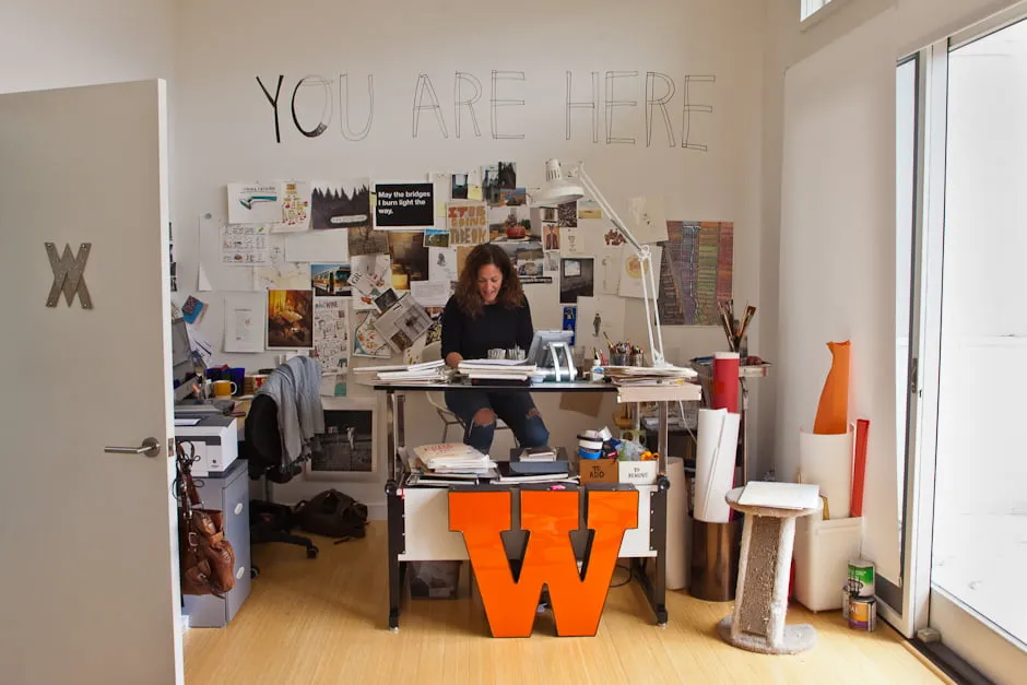 Make work. - myheartcreative A female artist works at a desk at the In The Make studio. Art supplies are scattered around, with drawings taped to the wall behind her.