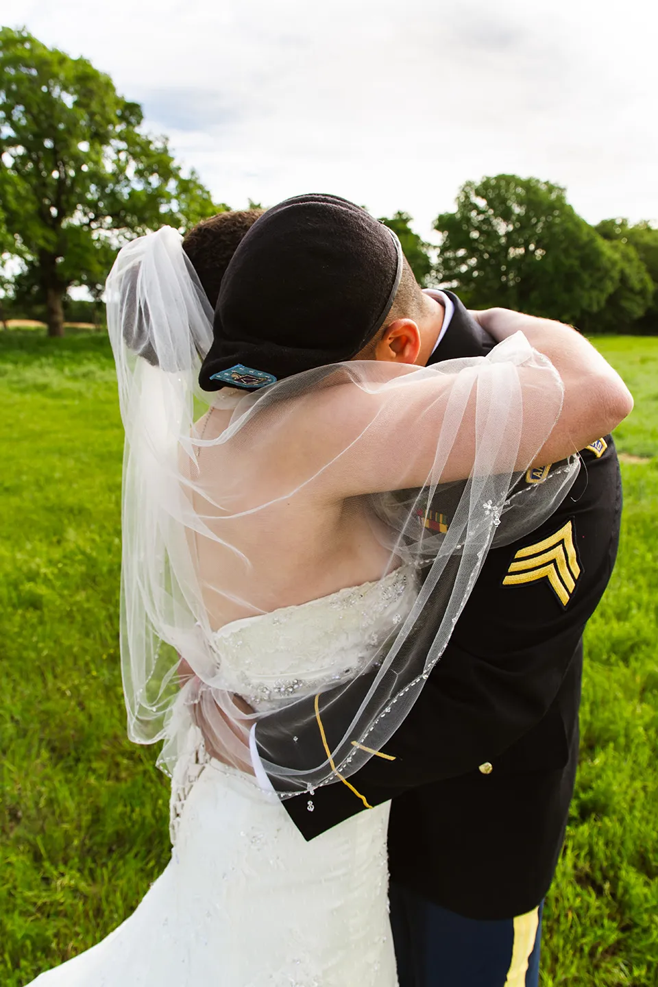 The groom hugs the bride and buries his face into her shoulder. They are standing in a field. 
