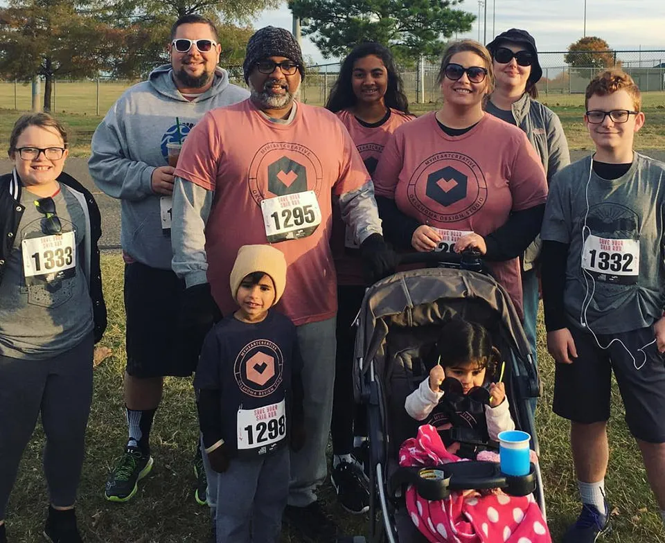 Sam, Amy, members of the studio team, and their children smile for a group photo at the Miles for Melanoma run. Everyone is wearing matching branded t-shirts. 