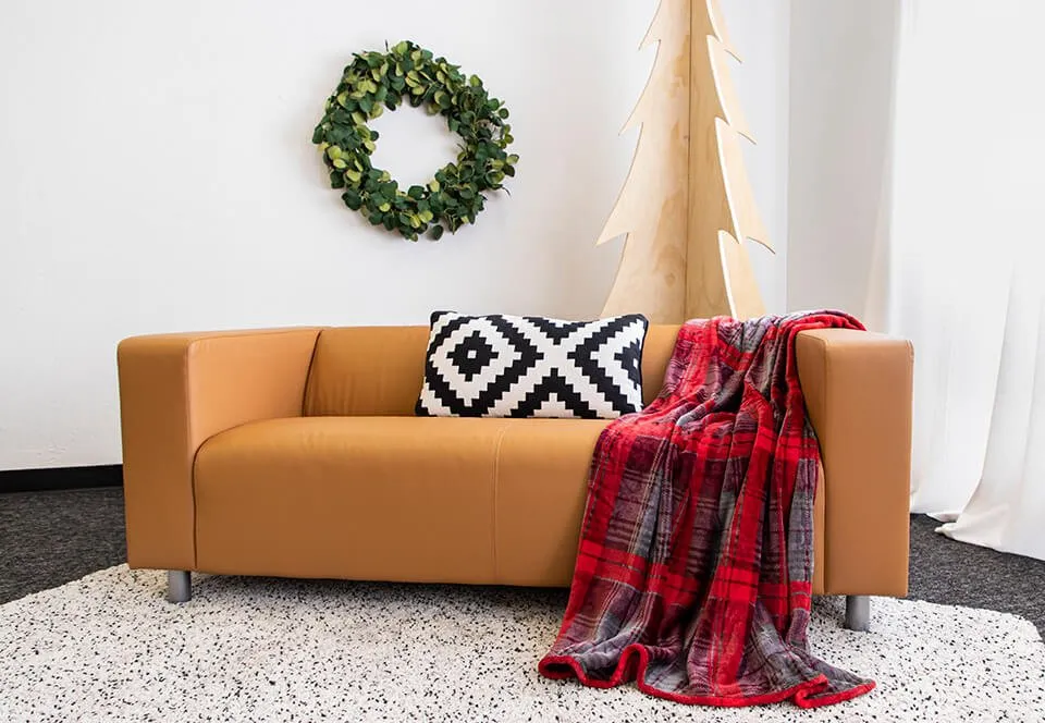Holiday Pictures - myheartcreative The set-up for a mini Christmas photoshoot at the myheartcreative studio. A tan couch sits on a tan rug with a black and white patterned throw pillow sitting in the center. There is a red and gray checkered throw blanket draped over the right side of the couch, with a greenery wreath hanging on the wall in the back. The set-up is complete with a wooden Christmas tree behind the couch.
