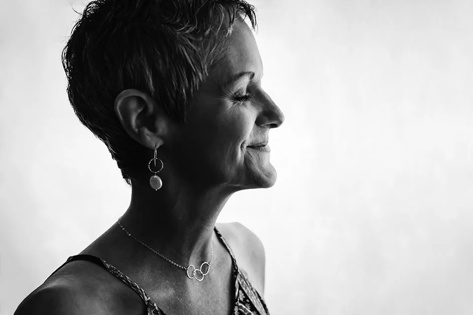 A black and white photo of a breast cancer survivor's side profile on a white background. She is closing her eyes and smiling softly. There is a dark shadow cast on part of her face to create a serious mood. 