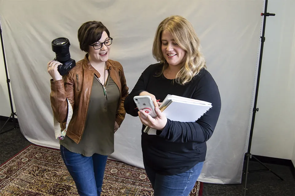 Looking Back on a Year of Web Design OKC - myheartcreative Amy shows Morgan a photo idea on her phone while holding a notebook. Morgan holds her camera and smiles. Both are standing in front of a white photoshoot backdrop.