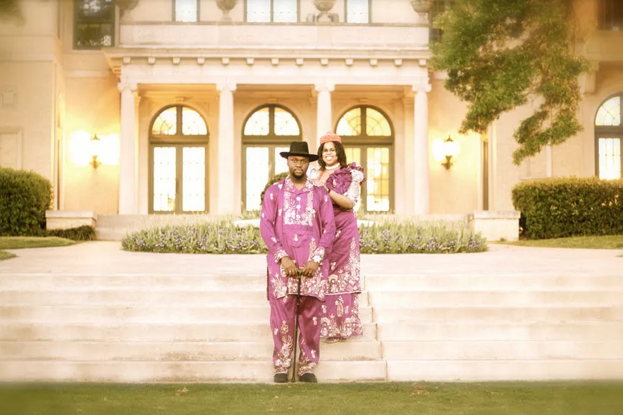 Mikki and Fred, the bride and groom, stand in front of their wedding venue in traditional purple African dress. 