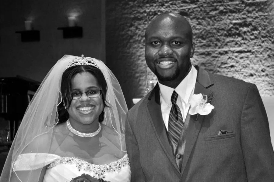 Mikki and Fred Oraene smile together with after their ceremony. Mikki wears a tiara, veil, and white wedding gown while Fred wears a suit and tie. 