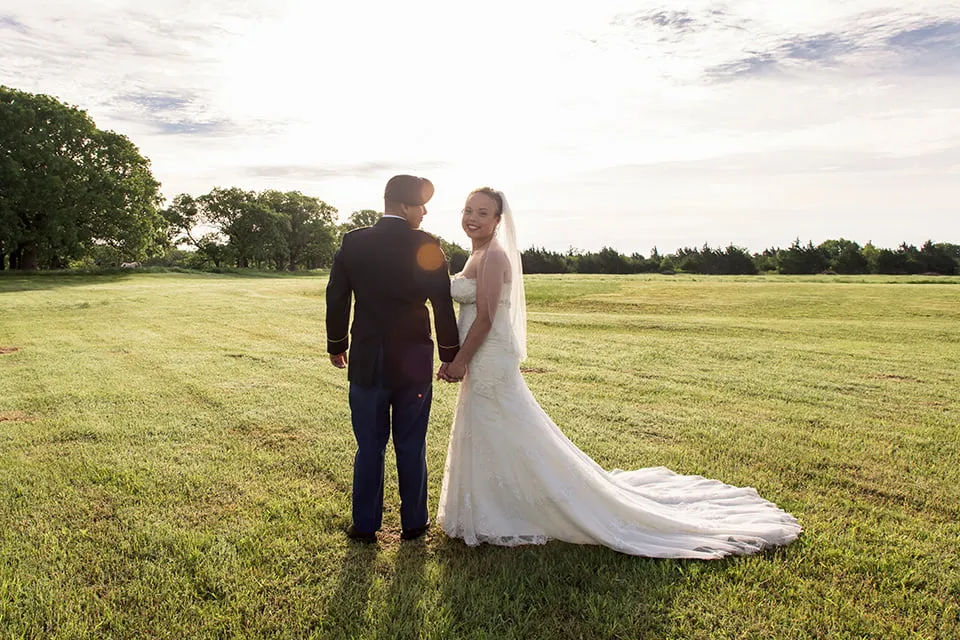 The bride and groom hold hands in a beautiful field. The bride looks back to the camera while the groom looks at her. 
