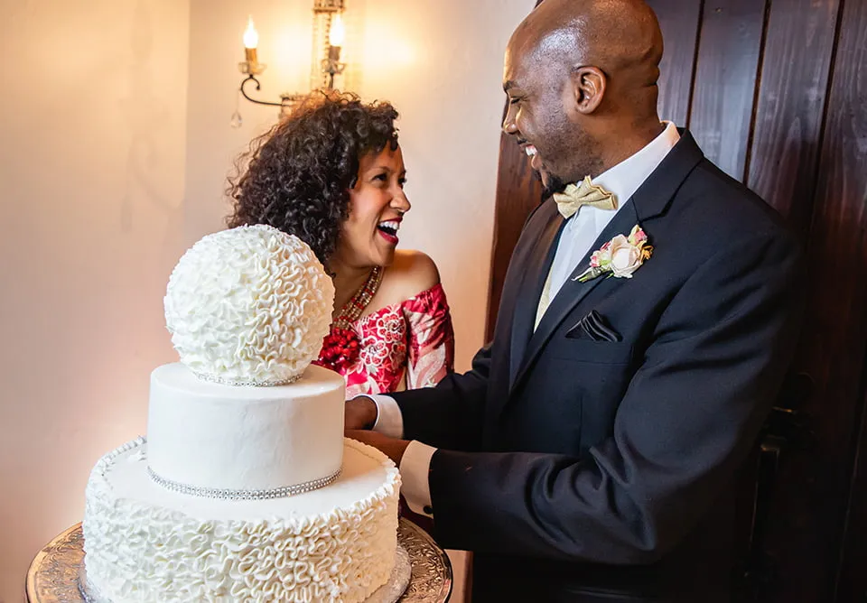 The bride wears a custom red patterned dress from Nigeria and the groom wears a black tuxedo. They smile and laugh as they cut a large white wedding cake. 