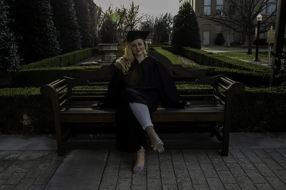 An image of a graduate sitting on a bench in her cap and gown. The image is dark and underexposed, so the subject is not easily seen. 