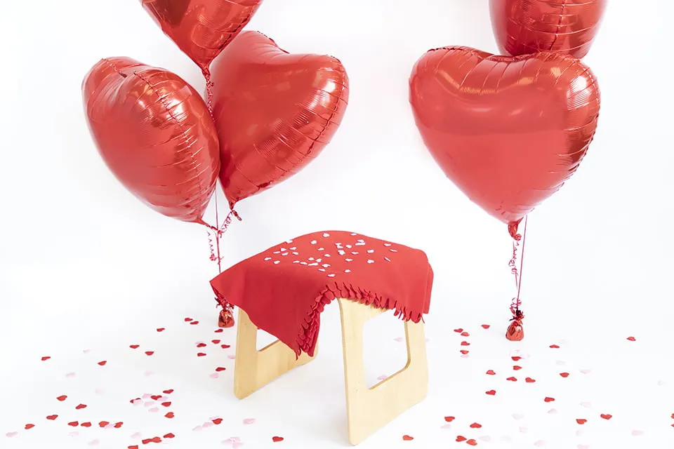 A set-up for a Valentine's Day mini session by a myheartcreative photographer OKC. The set-up includes a small stool with a red blanket, two bundles of red heart balloons on either side of the stool, red and pink confetti hearts scattered on the ground, all on a white background. 