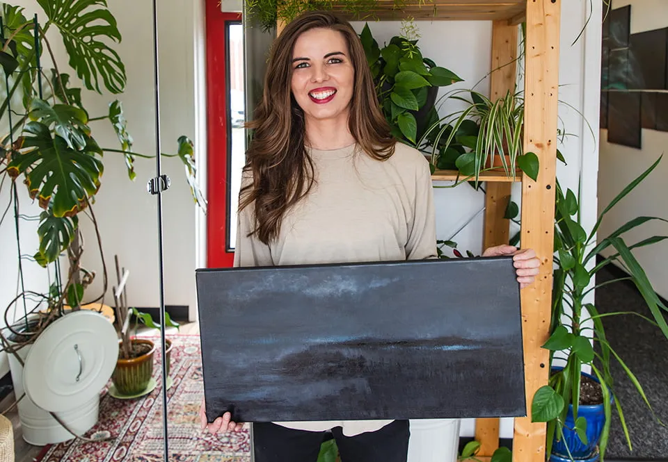 The Nature of Abstract - myheartcreative Artist Wendi O'Connor smiles while holding a black and blue abstract painting at the studio. She is standing in front of a wooden shelf with plants.