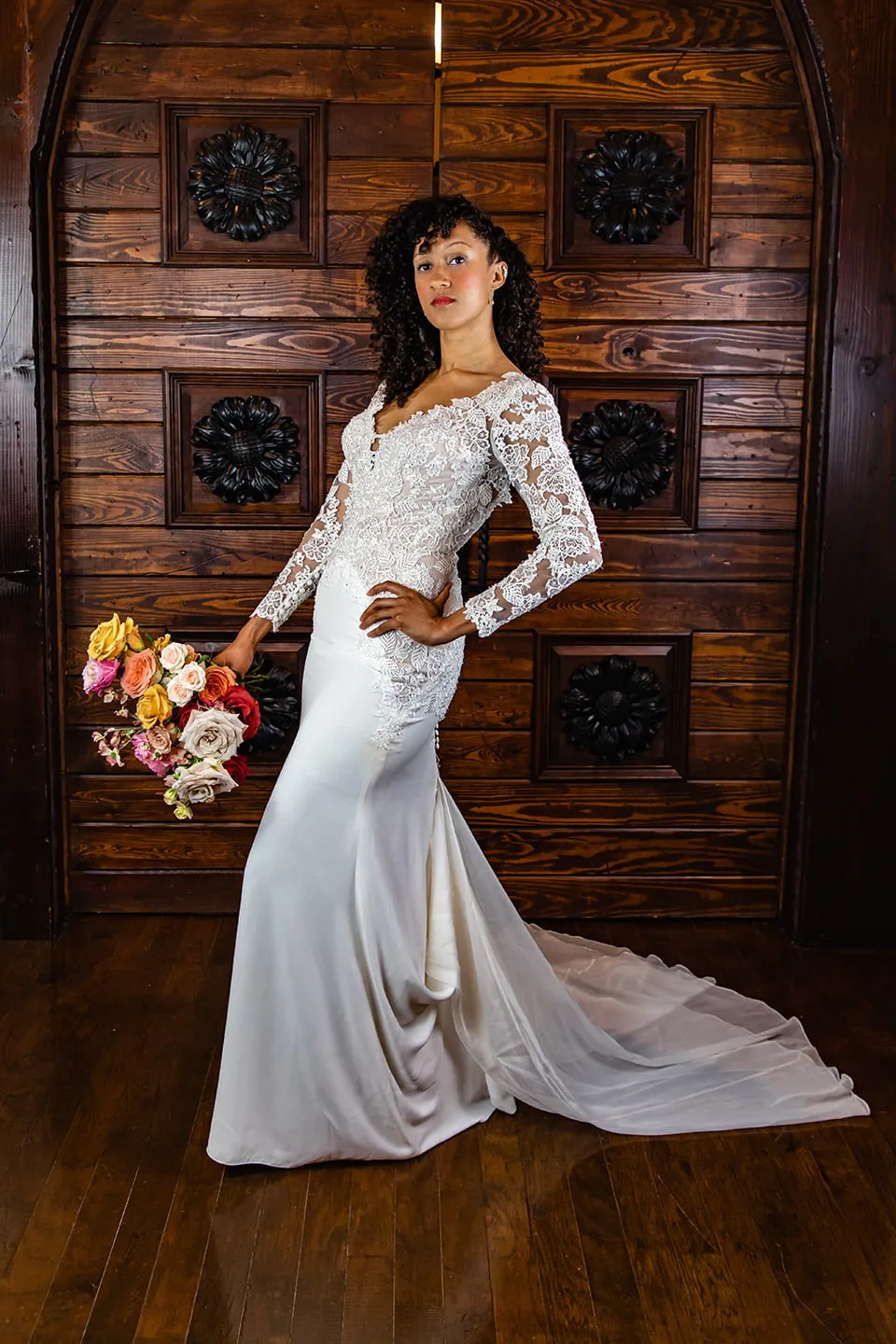 A bride wearing a white wedding gown with lace sleeves stands with her hand on her hip. She is in front of a large dark wooden door at Rose Briar Place.