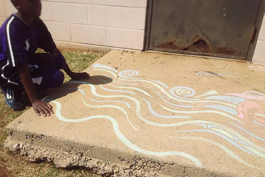 A young boy puts his hands on sidewalk pavement adorned with chalk art. 