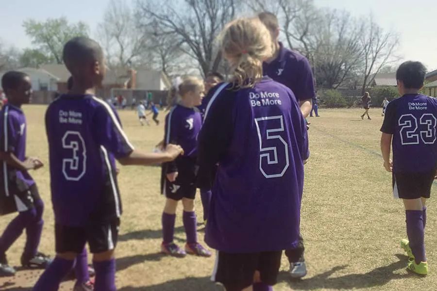 Young children gather around their coach. They are wearing purple jerseys that say, "Do More, Be More" on the back. 