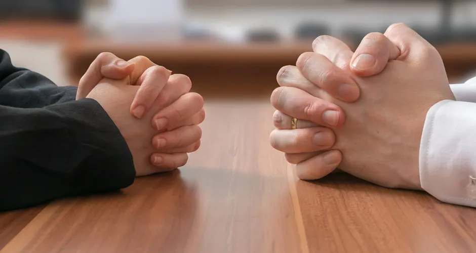 Two people sit across from one another at a wooden table with their hands clasped together. 