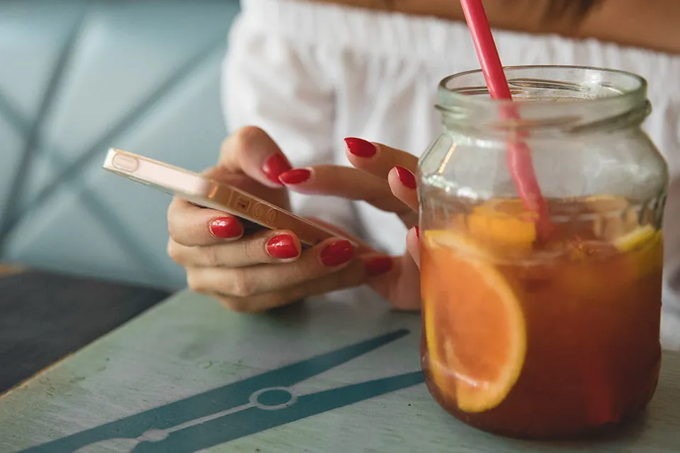 A woman types on her iPhone while sitting at a restaurant. Her fingernails are painted red. A mason jar filled with iced tea with oranges floating in it sits in front of her. 