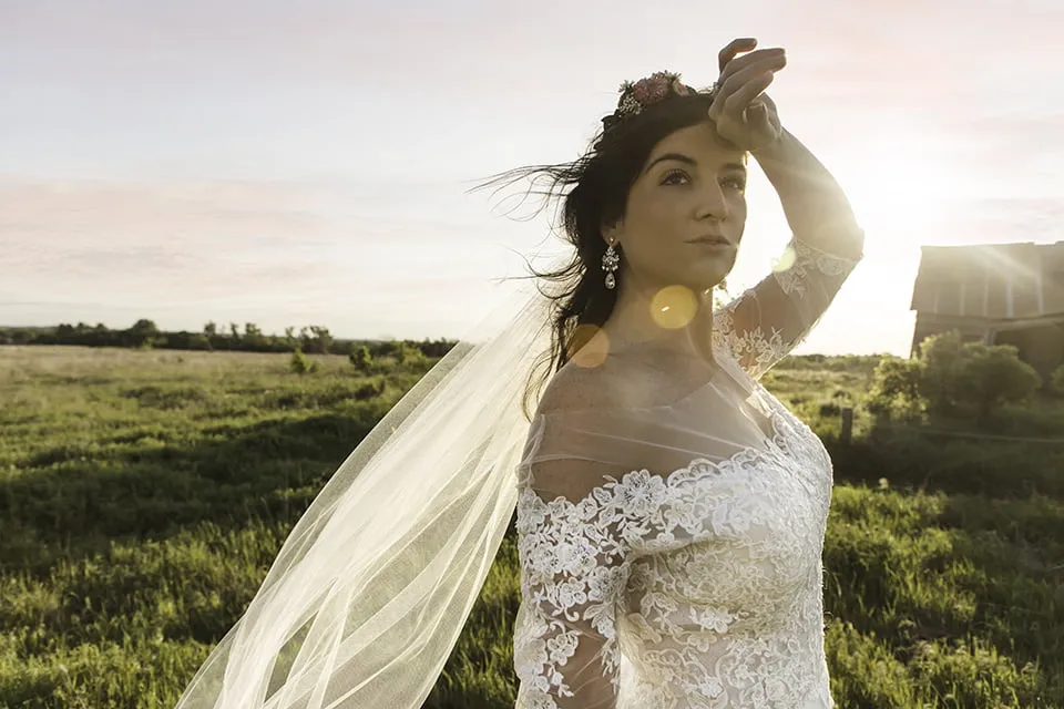 The bride wears a flower crown and holds her hand up to her head while posing in front of an old barn. 