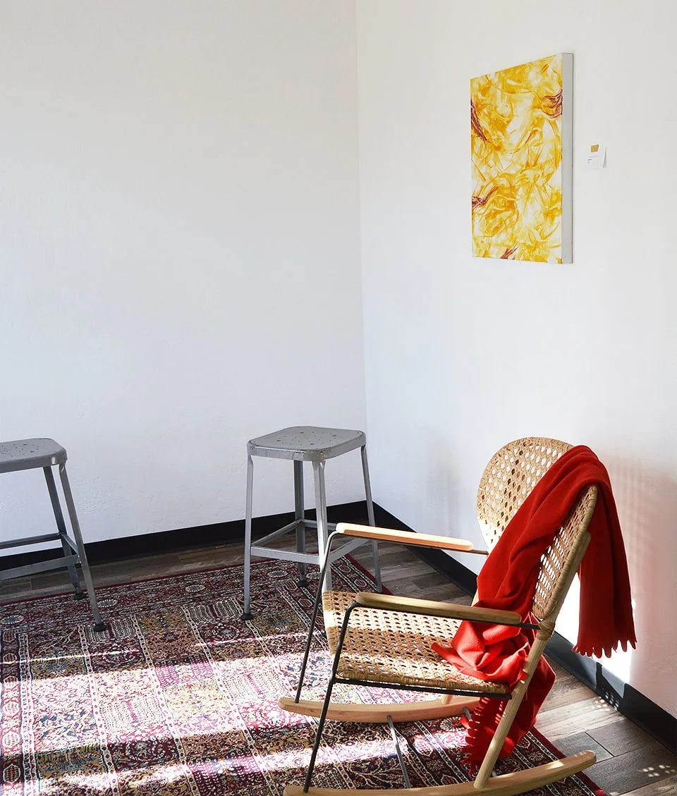 A corner of the studio with a yellow abstract painting by Theresa Hultberg. Two grey metal stools and a tan wicker chair surround the painting. 