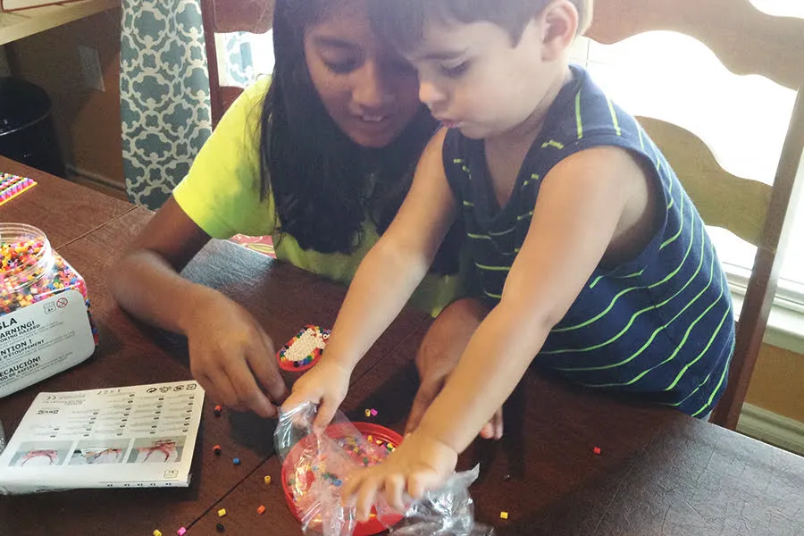 A young girl and her little brother sit at a wooden table and play with bead artwork. 