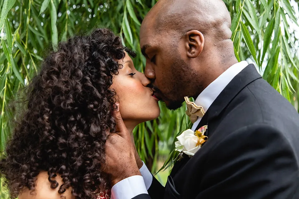 The groom grabs both sides of the bride's face and the two share a kiss in front of a weeping willow tree. 