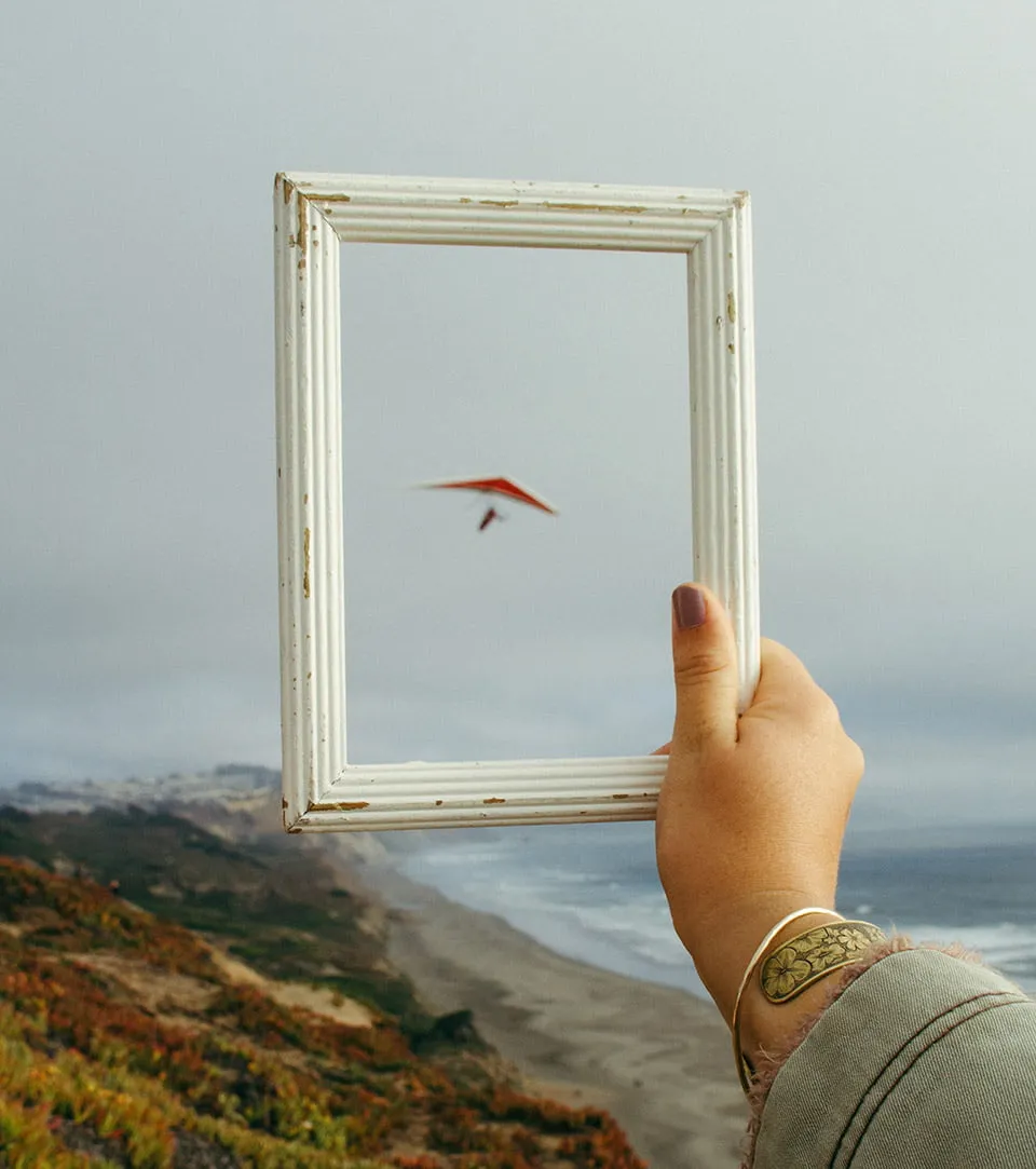 A woman holding a picture frame with a paraglider positioned in the middle of the empty frame. The frame and her hand are in focus; the paraglider is not. 