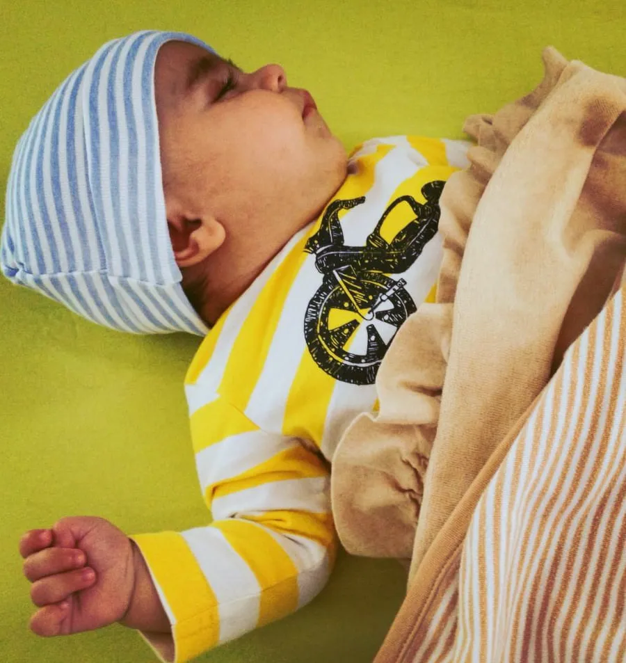 Baby Maximus sleeps on his back. He wear a blue and white striped hat and a yellow and white striped shirt with a motorcycle. 