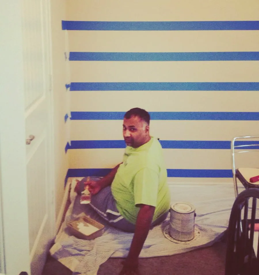 Sam sits on the floor as he paints the wall in Maximus's nursery. Rows of blue painters tape outline stripes on the wall. 