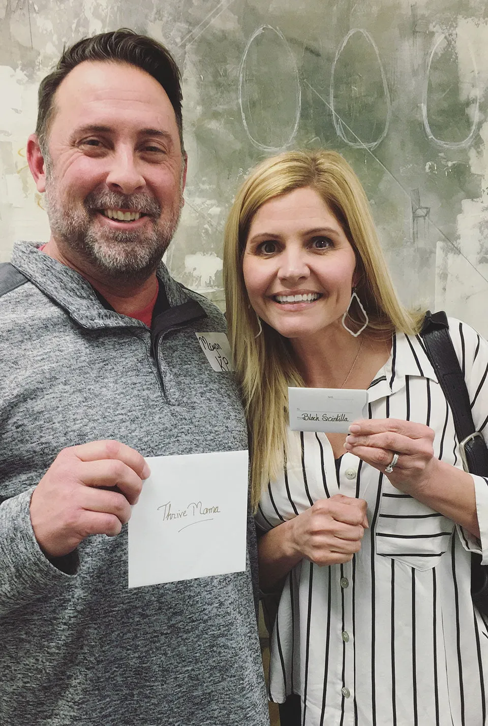 A woman holds up a gift card from the Black Scintilla that she won at the client appreciation party. She stands next to Mayor KP Westmoreland. He holds an envelope with "Thrive Mama" on the front. 