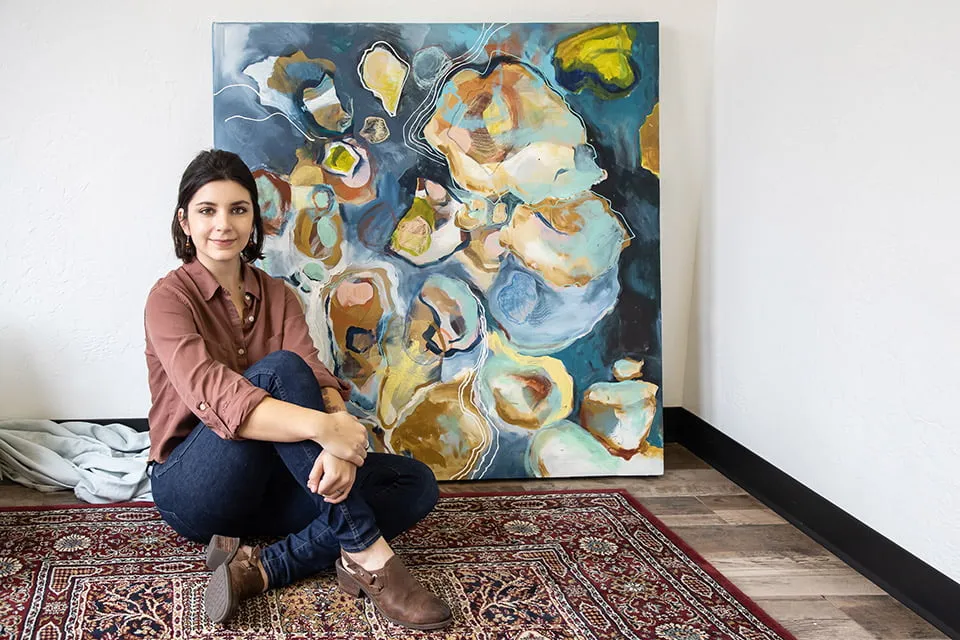 An Artistic View - myheartcreative Artist Mycah Higley sits on a patterned rug with her arms crossed around her legs. A large abstract painting sits behind her.