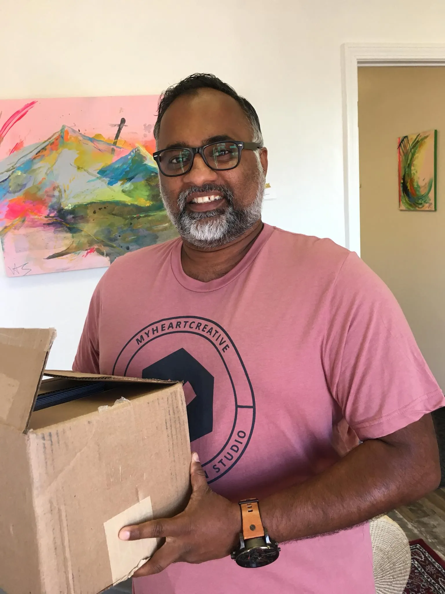 Sam, the Operations Director, smiles while holding a cardboard box. He is wearing a heathered red t-shirt with the myheartcreative logo on the front. 