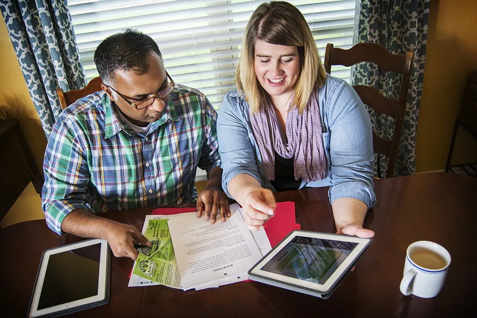 Sam and Amy work in their myheartcreative in-home studio at their kitchen table. Amy is holding an iPad while Sam views the screen. 