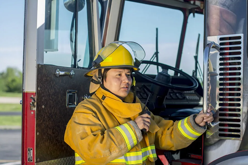 A firefighter stands in the doorway of a fire truck. He holds his call radio. 