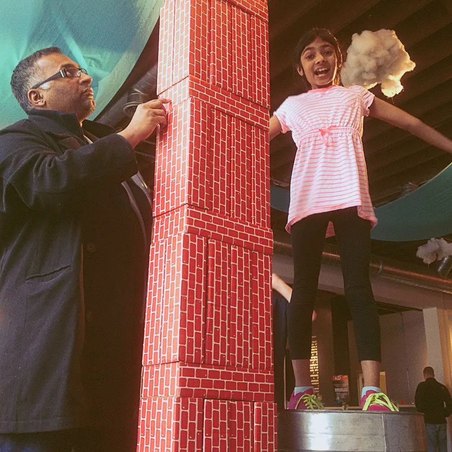 Sam builds a tall foam brick statue, while his daughter Kaitlynn stands on top of a metal platform next to him. 
