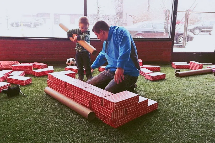 A young boy holds a long brown tube while his dad kneels next to him and builds a structure with foam bricks. 