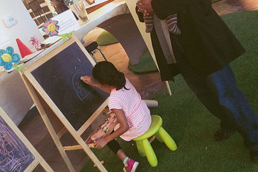 Kaitlynn sits at a small chalk board on a lime green stool and draws. Sam watches her. 