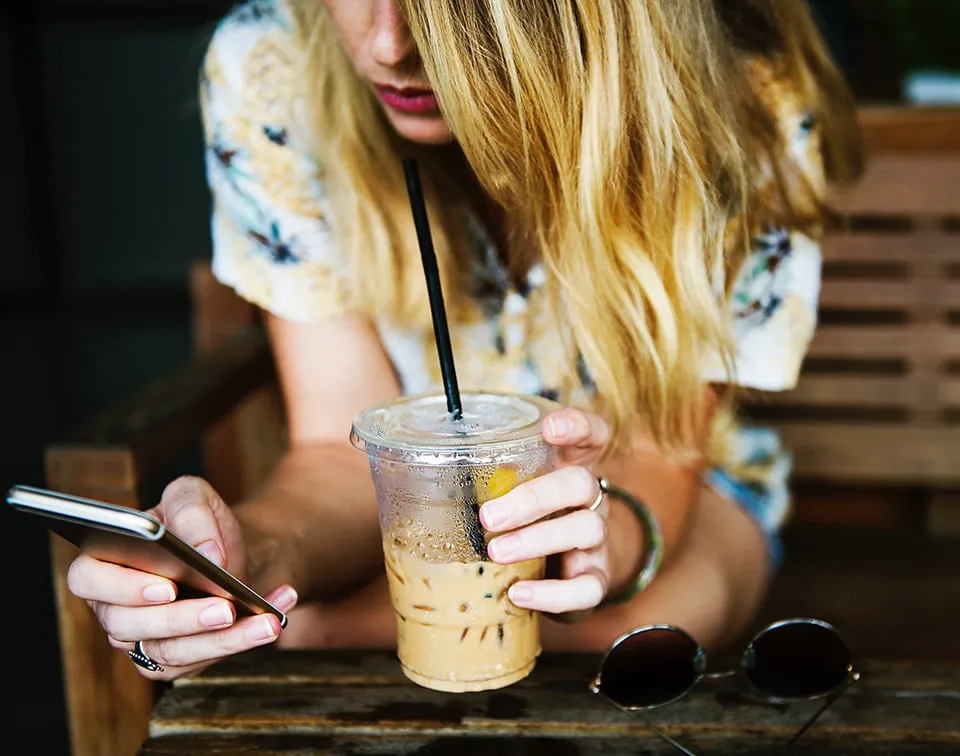A woman scrolls on her mobile phone while drinking an iced coffee. She is looking at a company's social media marketing ads. 