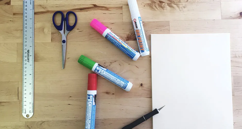 The supplies needed for a Christmas stencil project. A pair of scissors, a ruler, window markers, and exacto knife, and a white piece of paper sit on a table. 