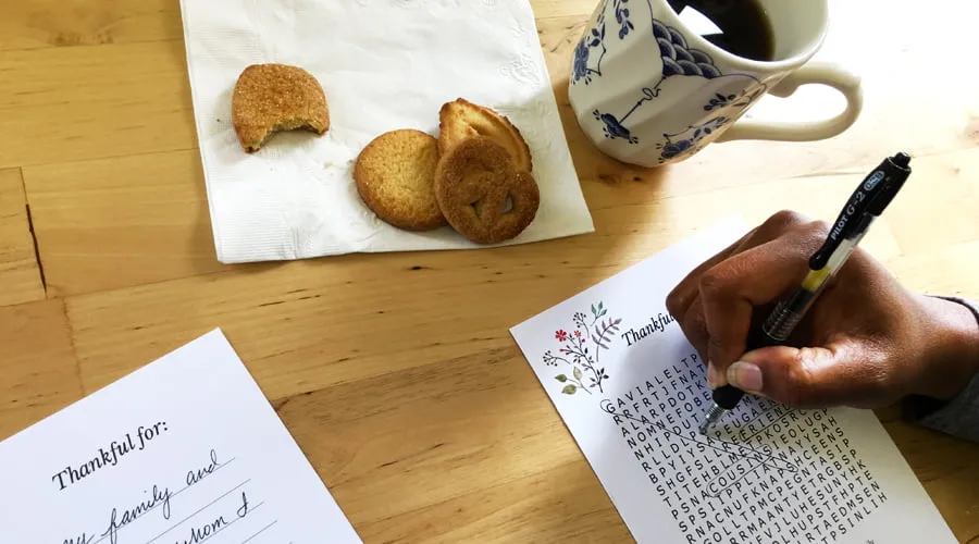 A hand circles a word on the Thanksgiving word search printable. The "Thankful For:" printable lays next to it, along with some cookies and a cup of coffee. 