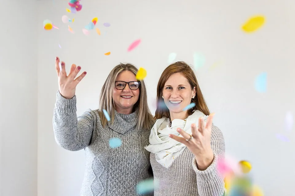 Our Clients Have Our Hearts - myheartcreative Two clients smile while throwing confetti in the air at the Client Appreciation Party. Both women are wearing tan sweaters.