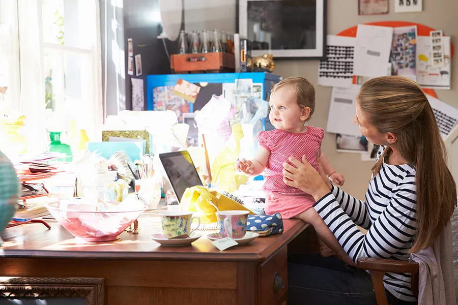 How to Be a Rockstar Remote Worker in a World of Employees - myheartcreative A mom talks to her baby daughter while sitting and working at her desk.